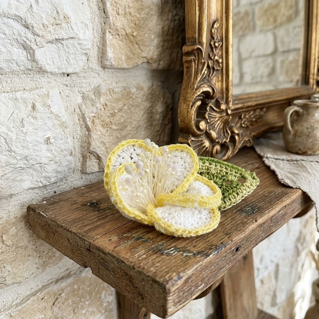 A handmade crocheted flower-shaped brooch in yellow and white, displayed on a rustic wooden shelf next to a green crocheted coaster, with a stone wall and an ornate gold mirror in the background.