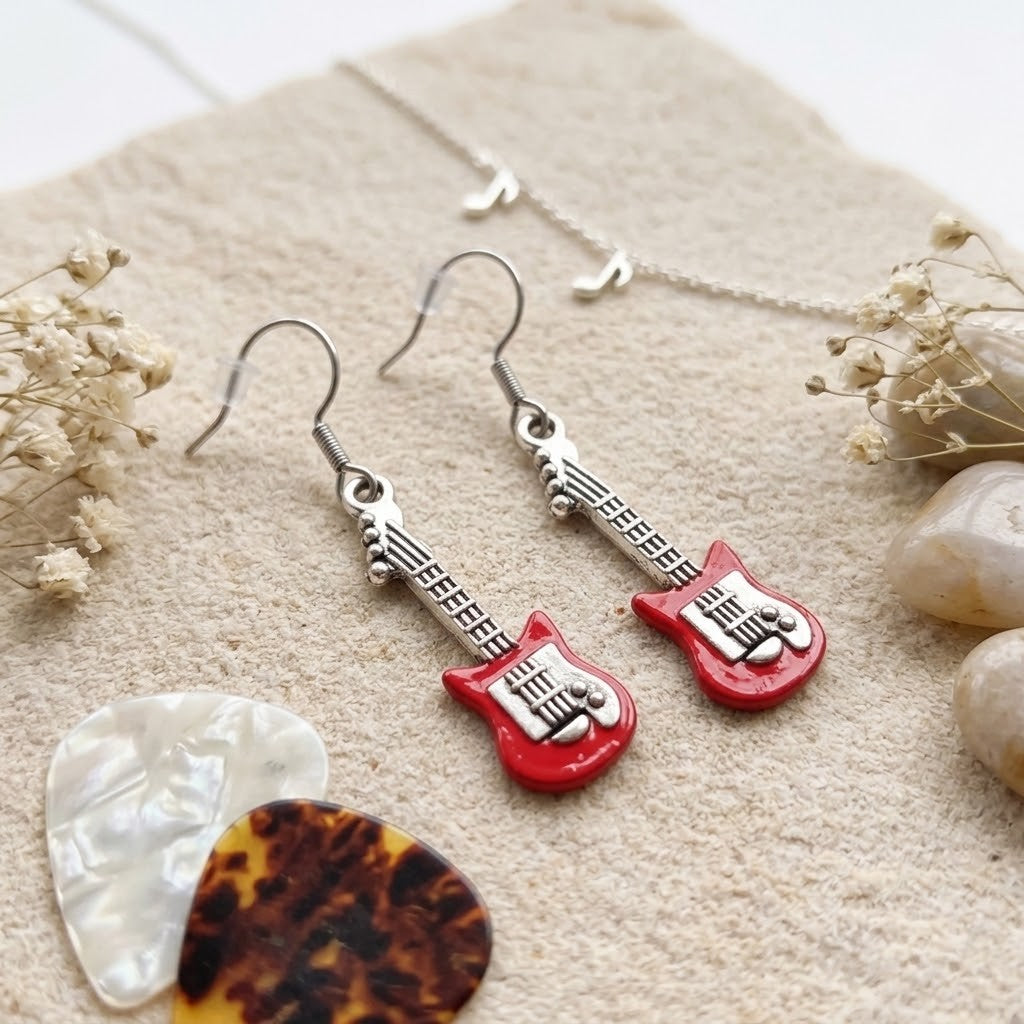 A pair of dangling earrings shaped like red electric guitars with silver-tone necks and hooks, displayed on a textured beige surface with dried flowers, stones, and a guitar pick.