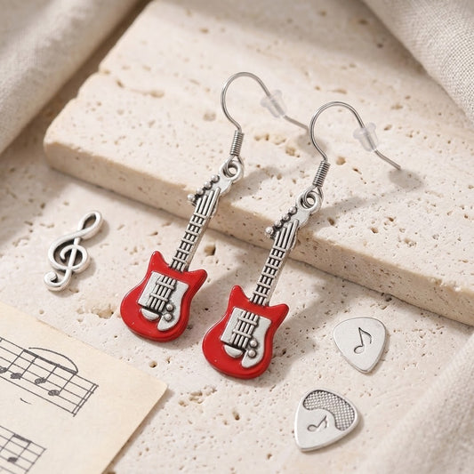 A pair of dangling earrings shaped like red electric guitars with silver-tone hooks and necks, displayed on a textured beige stone surface with small music-themed props including a treble clef, sheet music, and guitar pick charms.