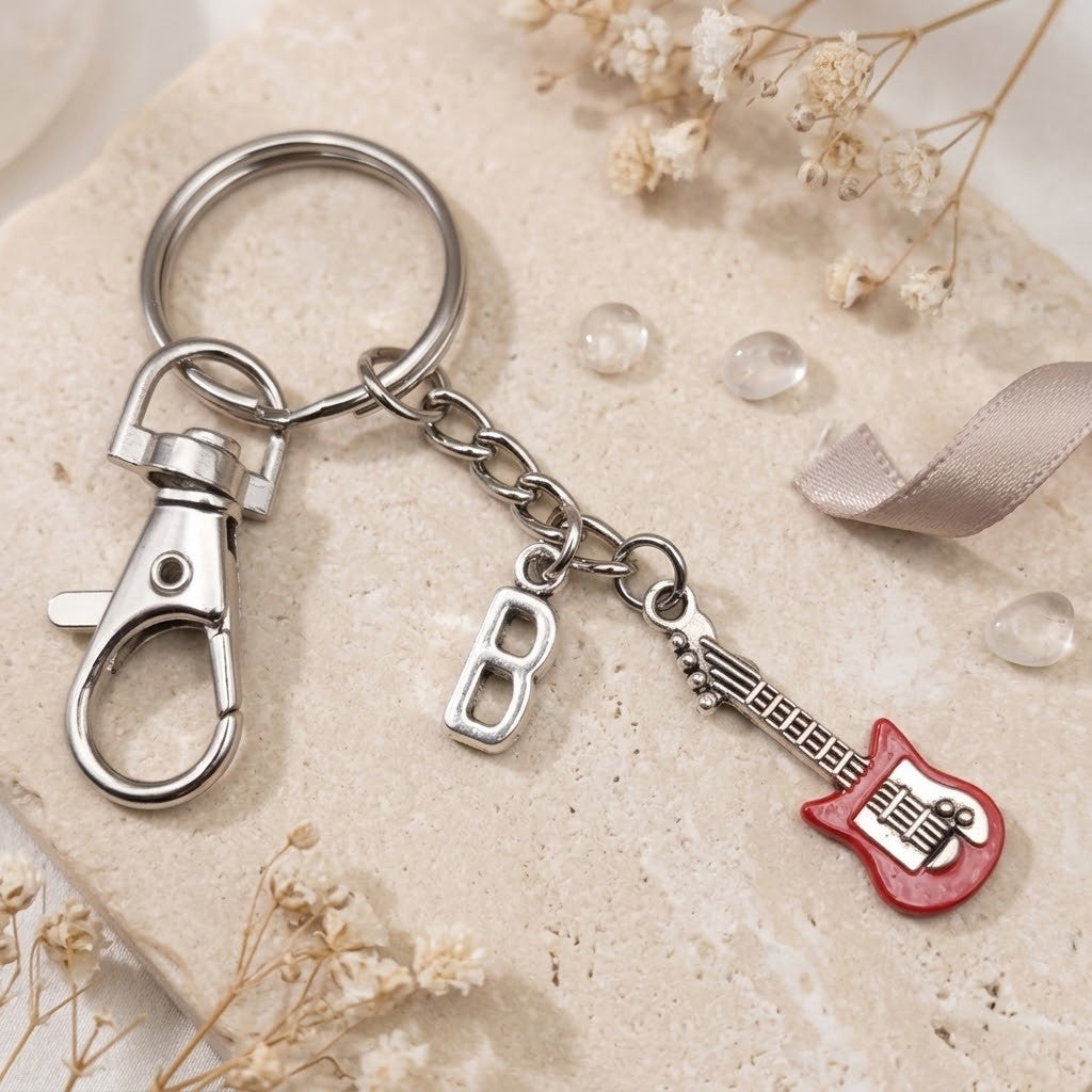 A silver metal keychain with a lobster clasp, a letter 'B' charm, and a red guitar-shaped charm, displayed on a textured beige stone with dried flowers and a ribbon.