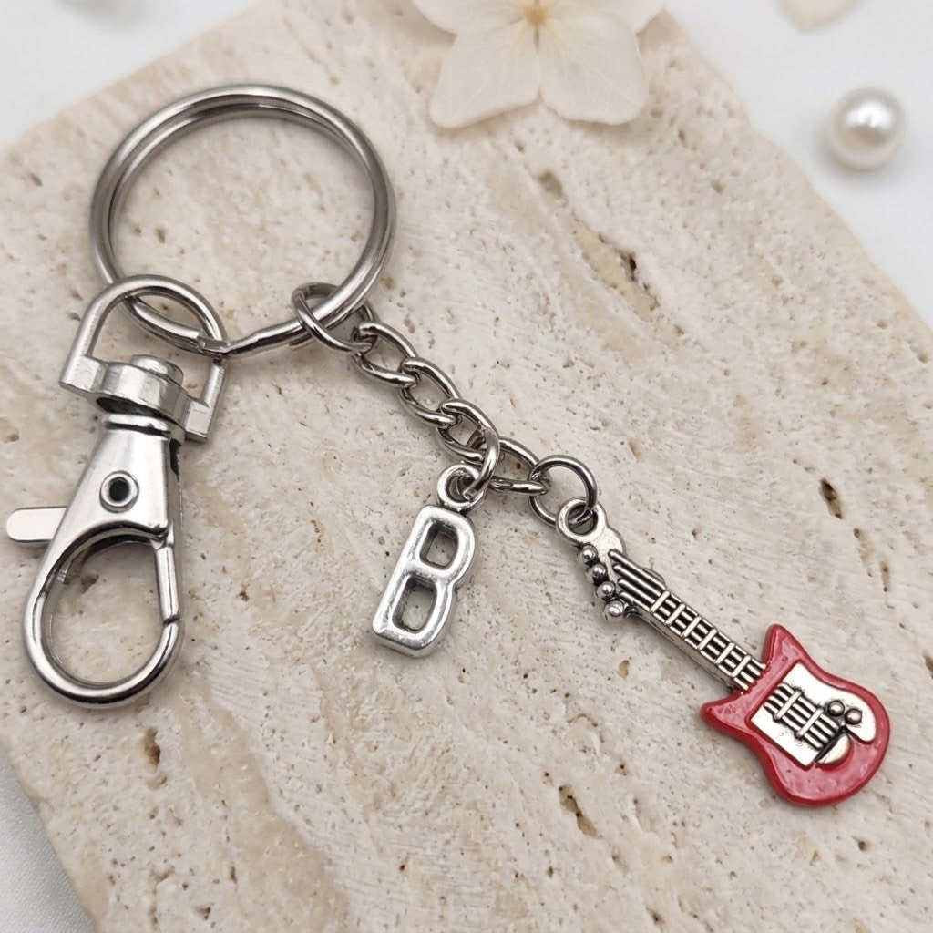 A silver metal keychain with a lobster clasp, a hanging letter 'B' charm, and a red electric guitar charm, displayed on a textured beige stone surface.