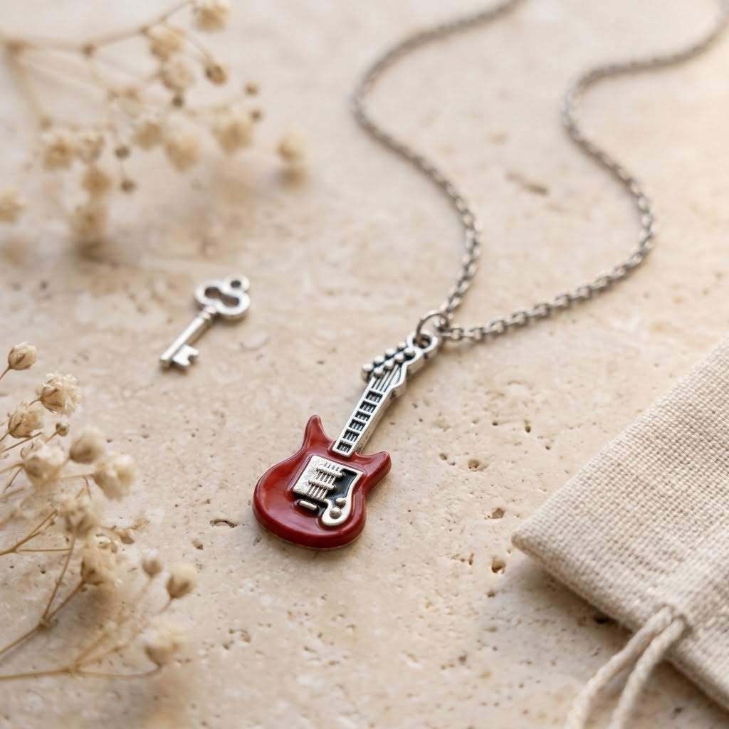 A silver-toned necklace with a guitar-shaped pendant in glossy red and silver, displayed on a textured beige stone surface with dried flowers and a small key prop.