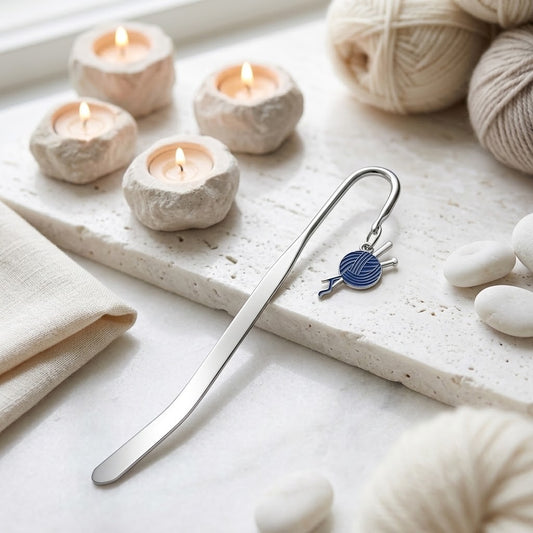 A silver metal bookmark shaped like knitting needles with a small blue and white yarn ball charm, resting on a light stone tray with candles, yarn balls, and white stones around it.