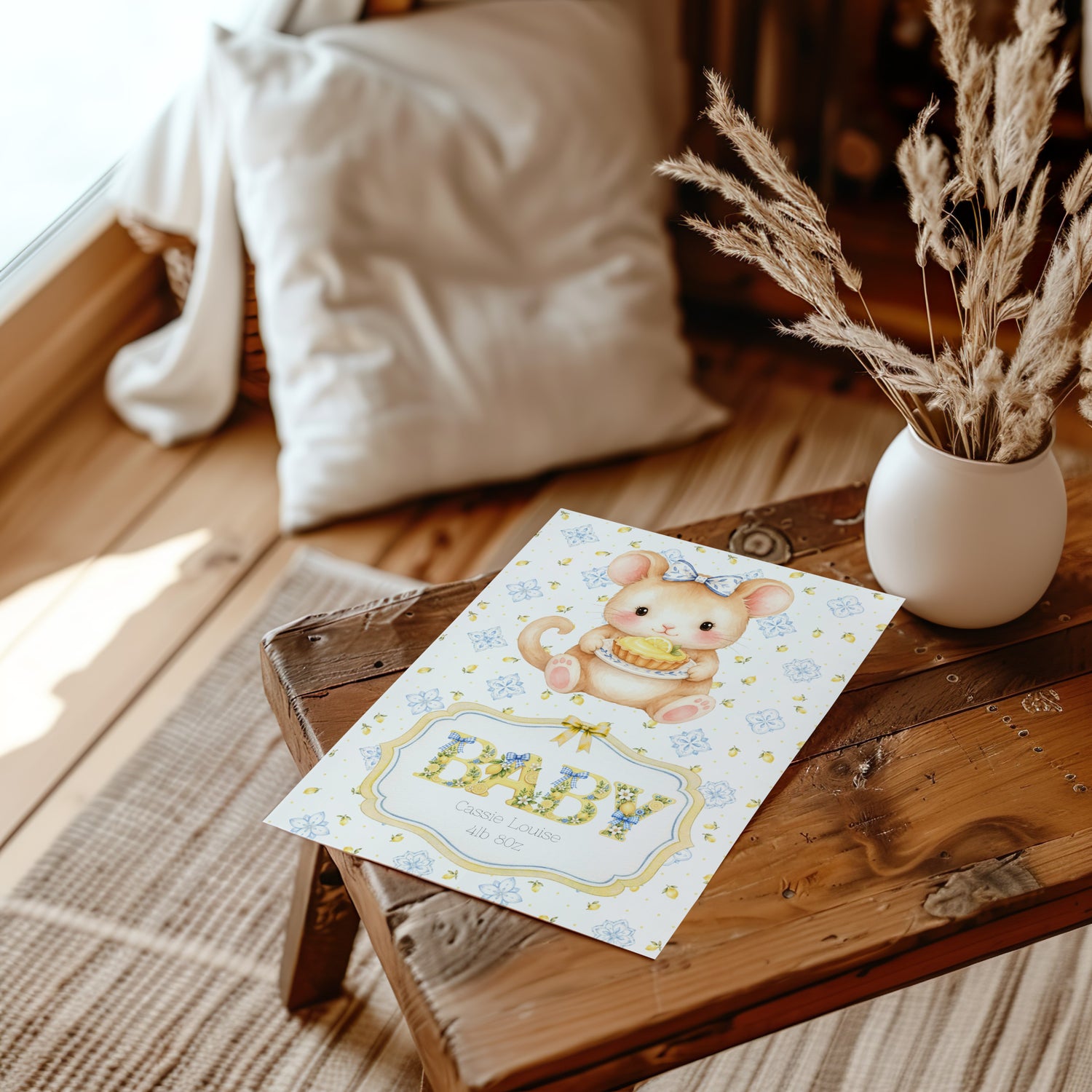 A newborn baby card featuring a cute cartoon bear holding a basket of yellow items, with the word 'BABY' in large decorative letters. The card is styled on a rustic wooden bench next to a white vase with dried grasses and a soft cream pillow in the background.