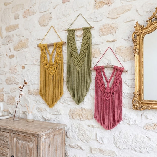 Three bohemian macrame wall hangings in mustard yellow, sage green, and dusty rose pink, displayed on a rustic white stone wall next to a gold-framed mirror.