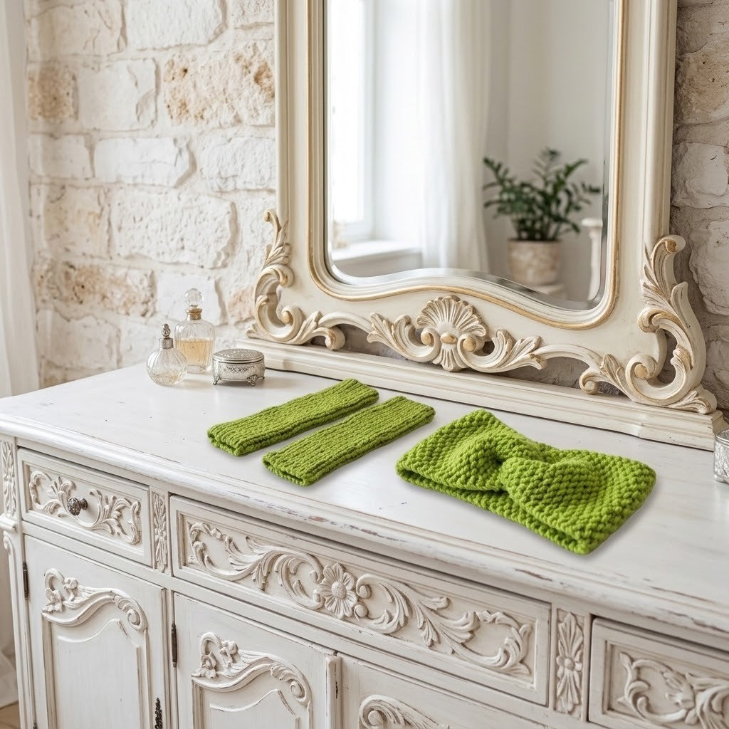 A set of bright green knit accessories, including a large knotted headband and two matching fingerless gloves, displayed on a distressed white wooden console table in front of an ornate cream mirror.