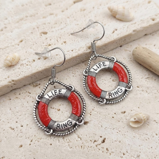 A pair of dangling earrings shaped like life rings, with red enamel-style sections, silver-toned rope-like borders, and the words “LIFE RING” engraved on the rings. They are photographed on a light stone surface with small seashells and a piece of driftwood as styled props.
