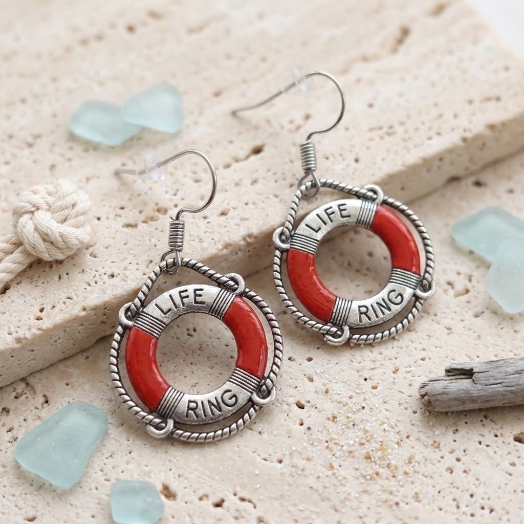 A pair of dangling earrings shaped like life buoys, with silver-toned metal wire frames, red enamel rings, and the words “LIFE” and “RING” engraved on the front. They are displayed on a light beige textured stone surface with small sea glass pieces and a rope knot as styled props.