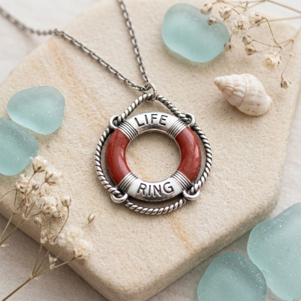 A silver-toned life preserver pendant necklace with a red-orange ring and the words 'LIFE' and 'RING' engraved on the sides, displayed on a stone slab with sea glass and dried flowers.