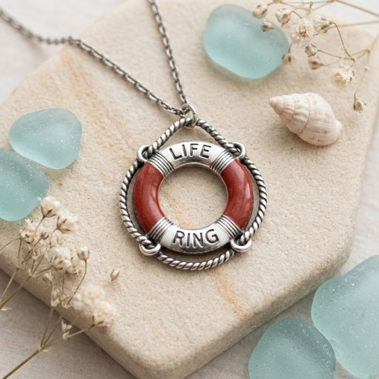 A silver-toned life preserver pendant necklace with a red-orange ring and the words 'LIFE' and 'RING' engraved on the sides, displayed on a stone slab with sea glass and dried flowers.