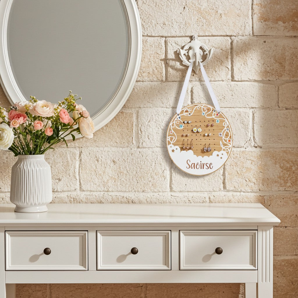 A personalised round wooden jewelry display board hanging from a white wall hook above a white console table. The board is printed with the name 'Saeirse' in script and decorated with white floral borders, holding several small earrings on white loops.