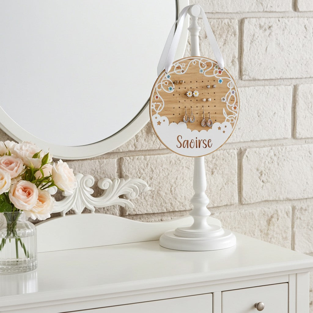 A personalised wooden jewellery display stand on a white pedestal base, decorated with a round wooden face, white lace cutout details, and a name at the bottom. It is styled on a white dresser next to a mirror and a vase of pink roses.