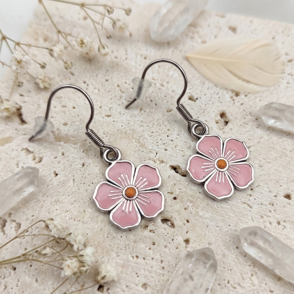 A pair of dangling flower earrings with pink enamel petals, silver-tone metal outlines, and small orange centers, displayed on a textured cream surface with dried flowers and clear crystal points.