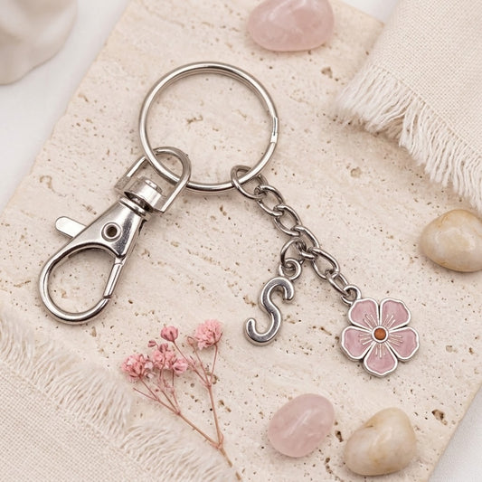 A silver metal keychain with a lobster clasp, split ring, and dangling charms including a pink flower and a pink letter S, styled on a textured beige fabric with small stones and dried flowers.