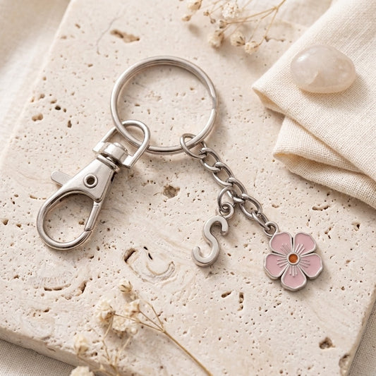 A silver metal keychain with a lobster clasp, a small chain, a lowercase letter charm, and a pink flower charm, styled on a textured beige stone tile with dried flowers and a folded cream fabric.