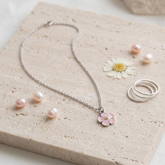 A silver chain necklace with a small pink flower pendant, styled on a beige stone slab with pink pearls, a daisy, and two silver rings.