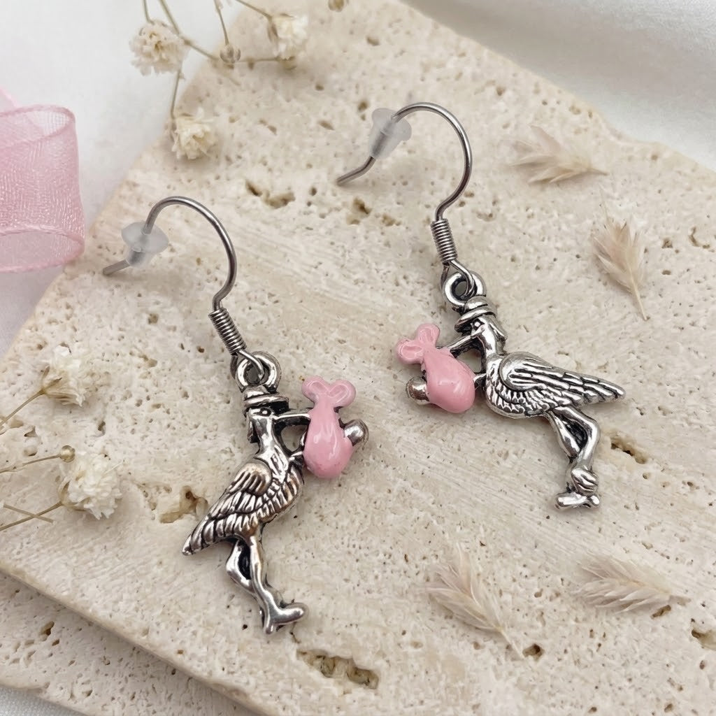 A pair of dangling earrings with silver-toned bird charms holding small pink envelopes, displayed on a textured beige stone surface with dried flowers and a pink ribbon.