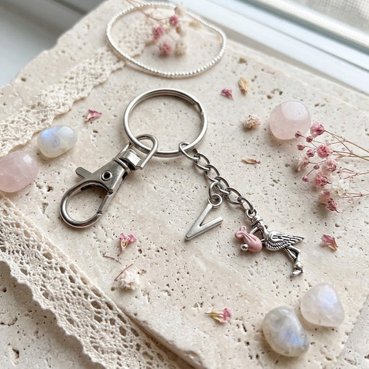 A silver-toned metal keychain with a lobster clasp, split ring, letter 'V' charm, and a small stork charm with pink accents, styled on a textured beige stone surface with lace, crystals, and dried flowers.