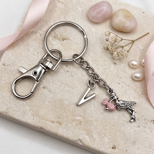 A silver-toned metal keychain with a lobster clasp, a split key ring, a dangling letter V charm, and a small stork charm with a pink accent, styled on a beige stone slab with pink ribbon and decorative stones.