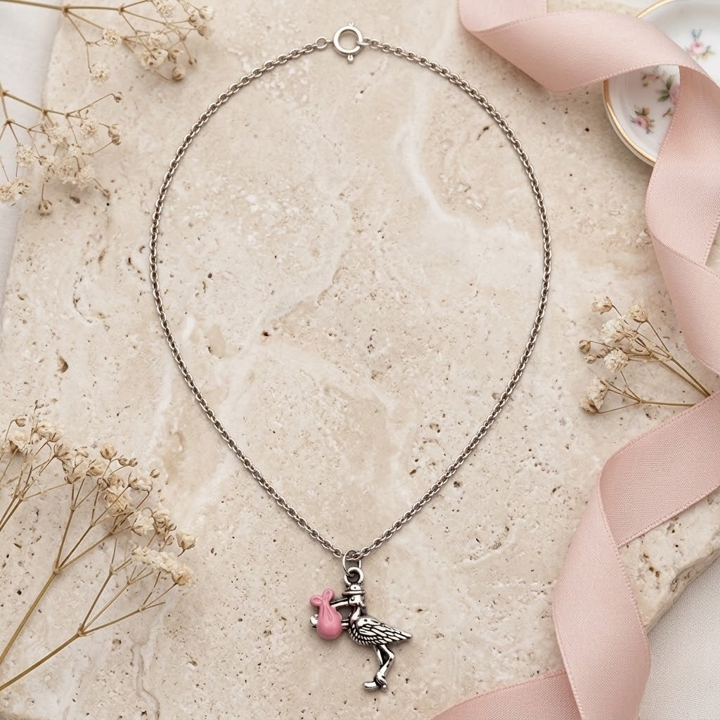 A silver-toned chain necklace with a stork charm, displayed on a beige stone surface with dried flowers and a pink ribbon.