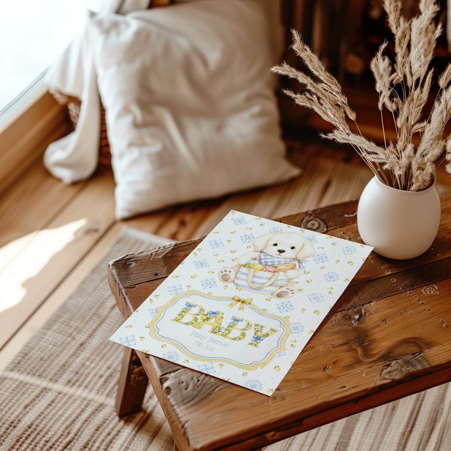 A baby shower card featuring a cute puppy in a striped shirt, with the word 'BABY' in large decorative letters. The card is styled on a rustic wooden bench next to a white vase with dried grasses and a soft cream pillow in the background.
