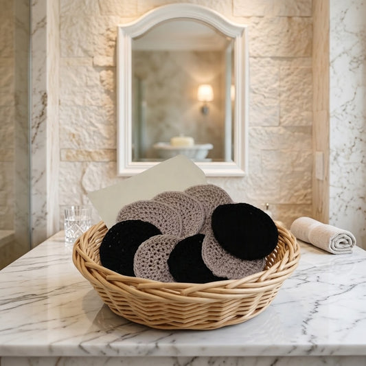 A woven wicker basket holding several round crocheted or knit face scrubbies in black and taupe, placed on a marble countertop in a bathroom setting with a mirror and stone wall behind it.