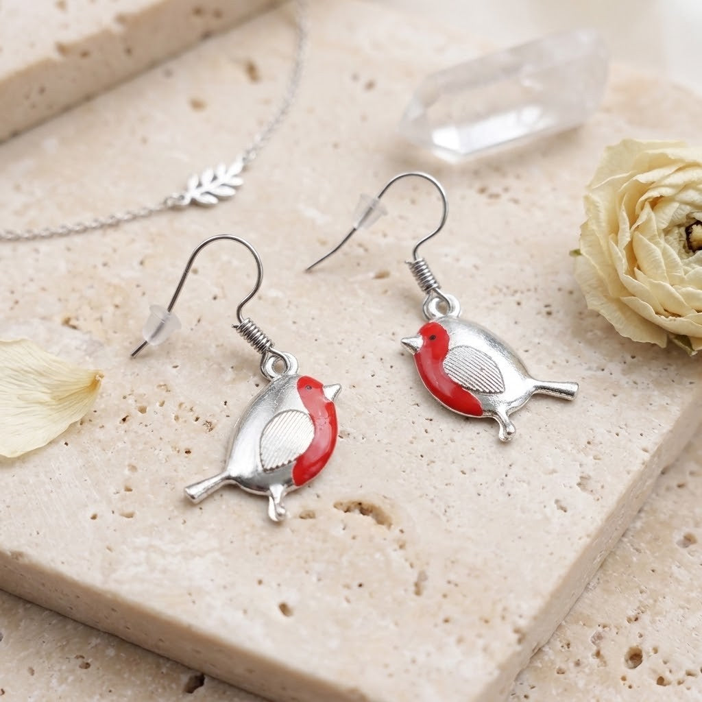 Silver bird drop earrings with red and white enamel details, displayed on a beige stone slab with a dried rose, crystal, and a silver necklace in the background.