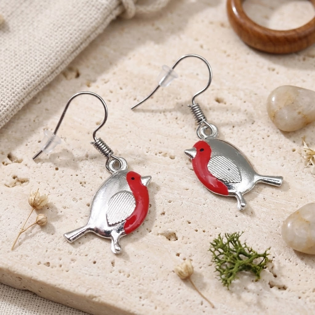 A pair of silver bird-shaped drop earrings with red enamel accents, displayed on a textured beige stone surface with dried flowers, small stones, and a burlap fabric in the background.