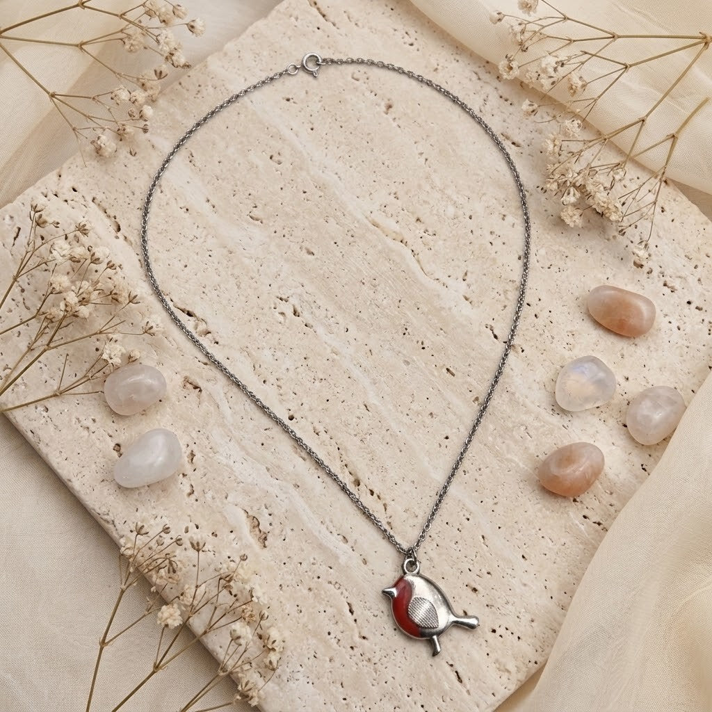 A silver-toned bird pendant necklace with a red enamel chest detail, displayed on a textured beige stone slab with dried flowers and polished stones.