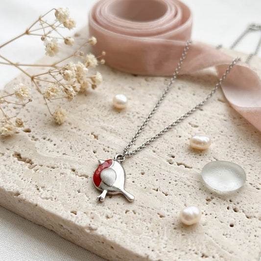 A silver-toned necklace with a bird pendant resting on a textured beige stone slab. The pendant features a red accent on the side.