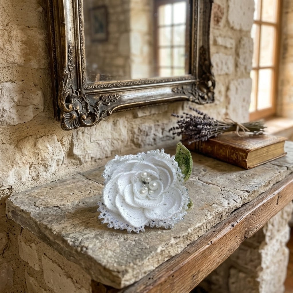 A white crocheted fabric rose brooch with pearl-like centre details, displayed on a rustic stone shelf next to an antique book and dried lavender under an ornate mirror.