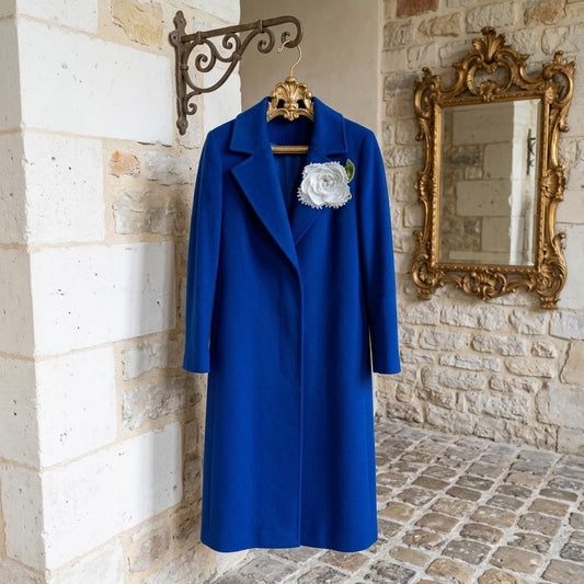 A vibrant royal blue wool coat hanging on a decorative gold hanger against a rustic stone wall, featuring a white fabric rose brooch on the left lapel.