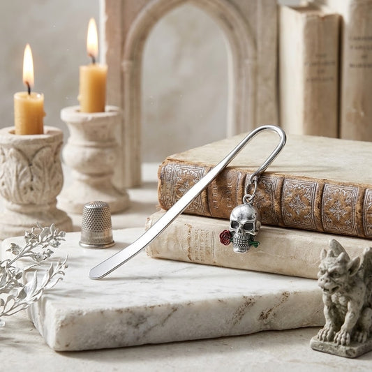 A silver metal bookmark shaped like a skull with small red and green gemstone accents, resting on stacked antique-style books on a marble slab. The scene is styled with lit candles, a thimble, a small gargoyle figurine, and a blurred gothic arch in the background.
