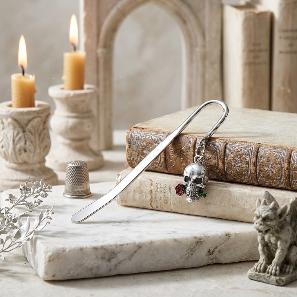 A silver metal bookmark shaped like a skull with small red and green gemstone accents, resting on stacked antique-style books on a marble slab. The scene is styled with lit candles, a thimble, a small gargoyle figurine, and a blurred gothic arch in the background.