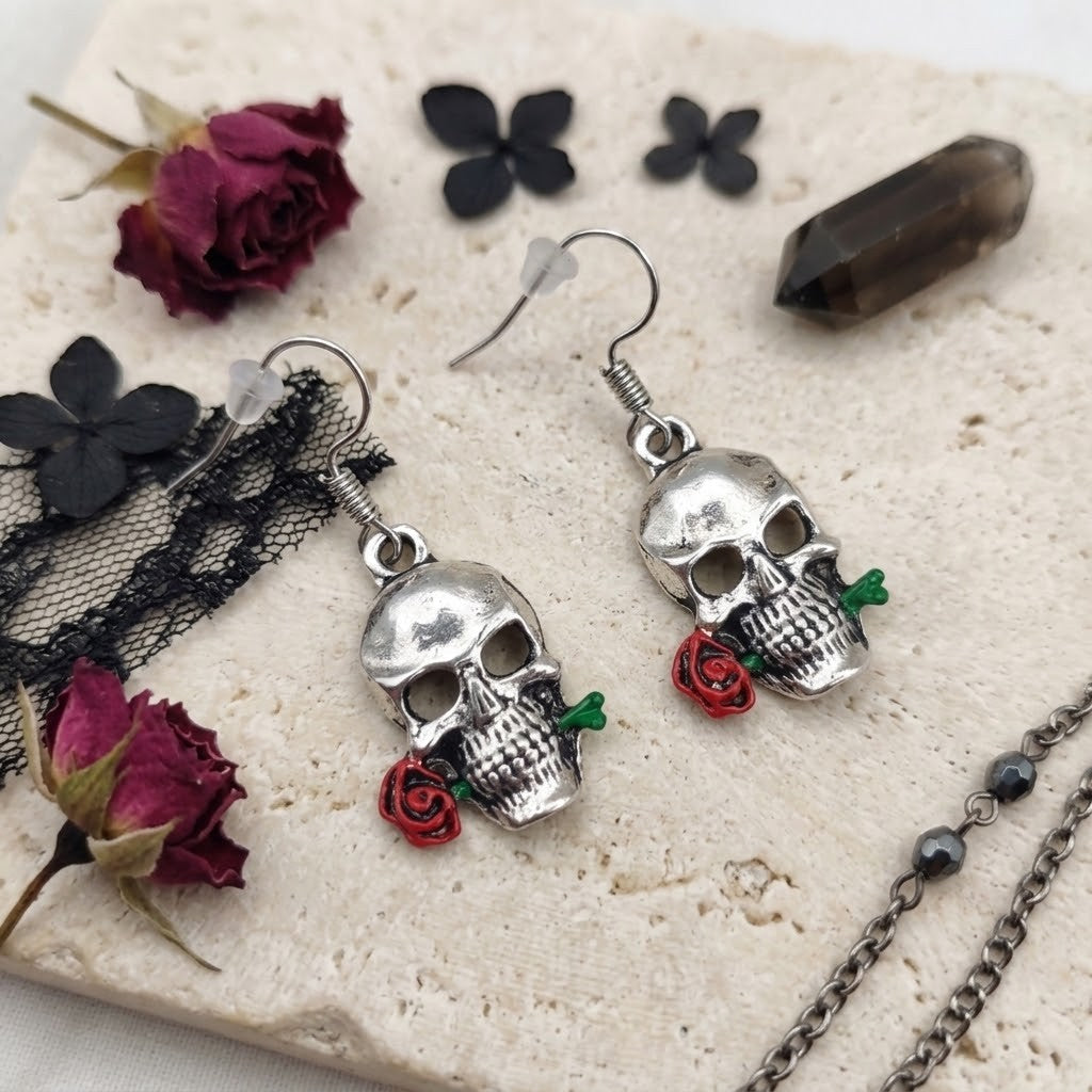 A pair of silver skull drop earrings with small red rose charms and green stems, displayed on a textured cream stone with dried roses, black lace, and a dark crystal.