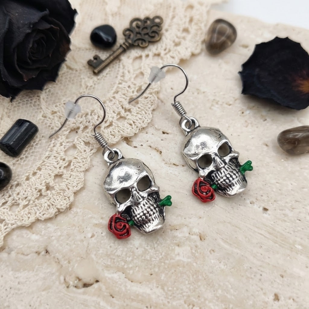 A pair of silver skull dangle earrings with small red rose charms and green stems, displayed on a textured beige surface with lace, dried black roses, and decorative stones.