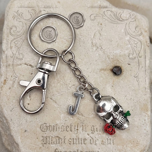 A silver-toned metal keychain featuring a lobster clasp, a split key ring, a hanging chain, a lowercase letter 'J' charm, and a skull charm holding a small red rose, displayed on a beige stone tile.