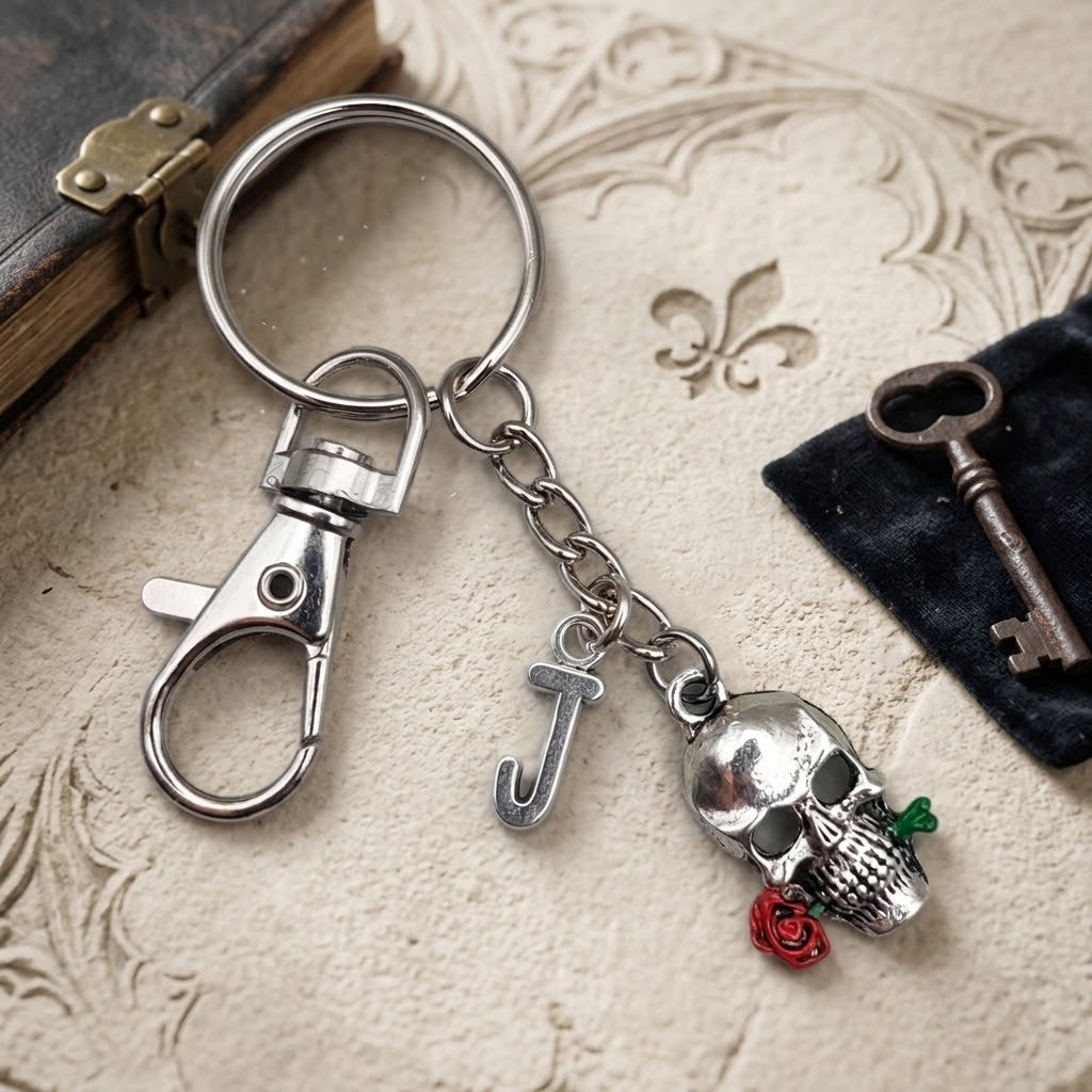 A silver-toned metal keychain with a lobster clasp, a letter 'J' charm, and a skull charm holding a red rose, displayed on a textured beige surface with vintage props.