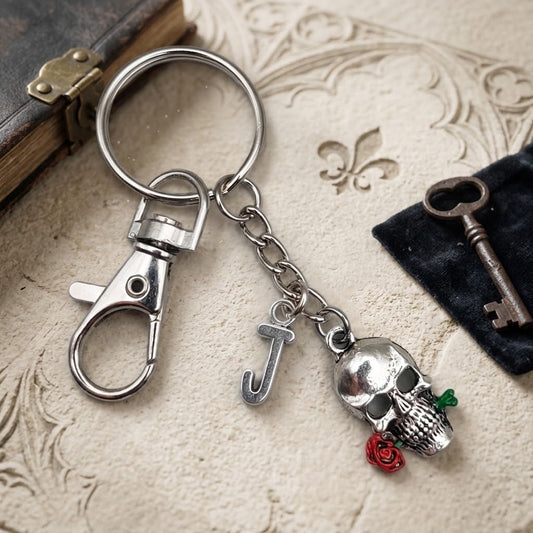 A silver-toned metal keychain with a lobster clasp, a letter 'J' charm, and a skull charm holding a red rose, displayed on a textured beige surface with vintage props.