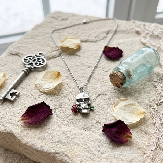 A silver-toned skull pendant necklace with a small red rose accent, displayed on a textured beige stone surface with dried flower petals, a vintage key, and a small glass vial.