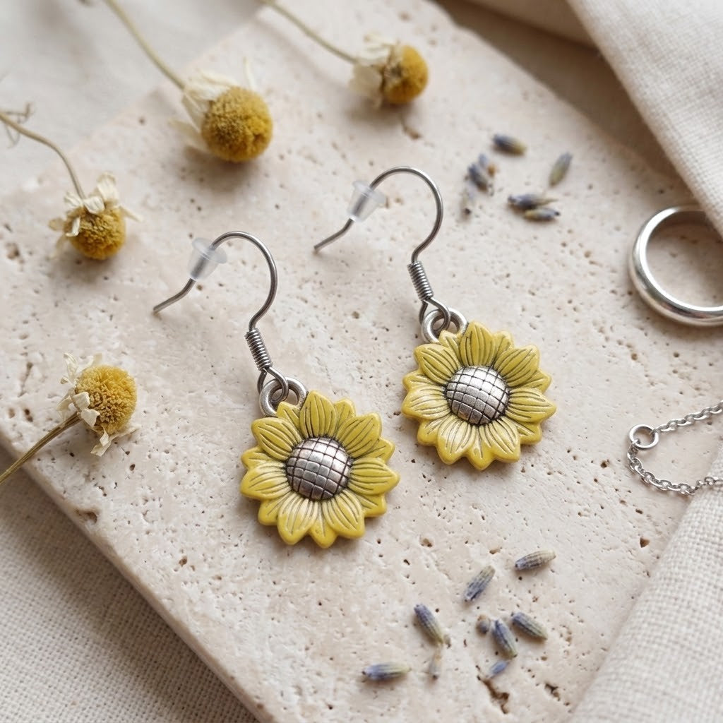 A pair of dangling sunflower earrings with yellow petals and textured silver centres, displayed on a beige stone slab with dried flowers and lavender.