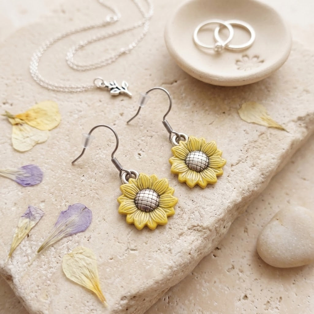A pair of dangling sunflower earrings with yellow petals and silver centers, displayed on a beige stone surface with dried flower petals, a necklace, and two rings in a small dish.