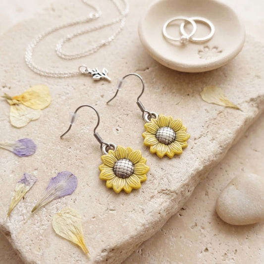 A pair of dangling sunflower earrings with yellow petals and silver centers, displayed on a beige stone surface with dried flower petals, a necklace, and two rings in a small dish.