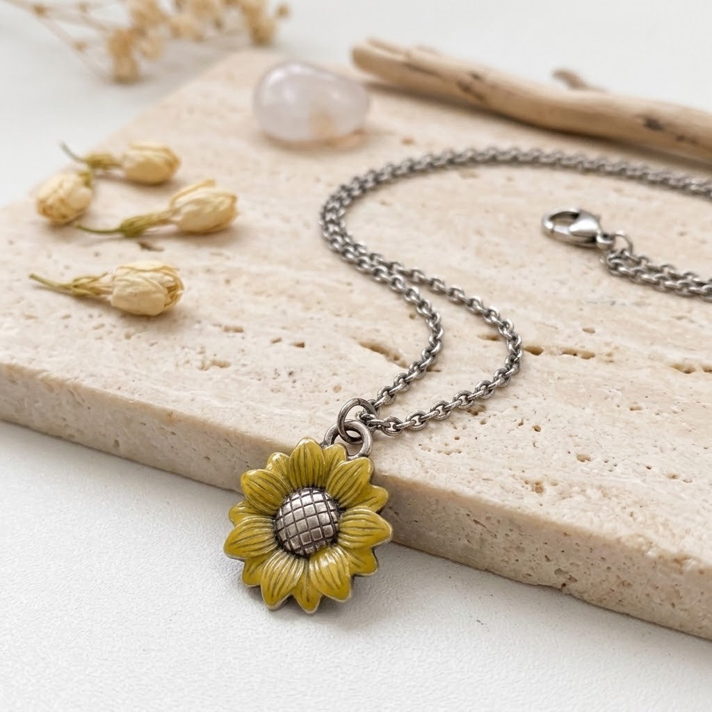 A silver-toned chain necklace with a detailed sunflower pendant, styled on a beige stone slab with dried flowers and a crystal.