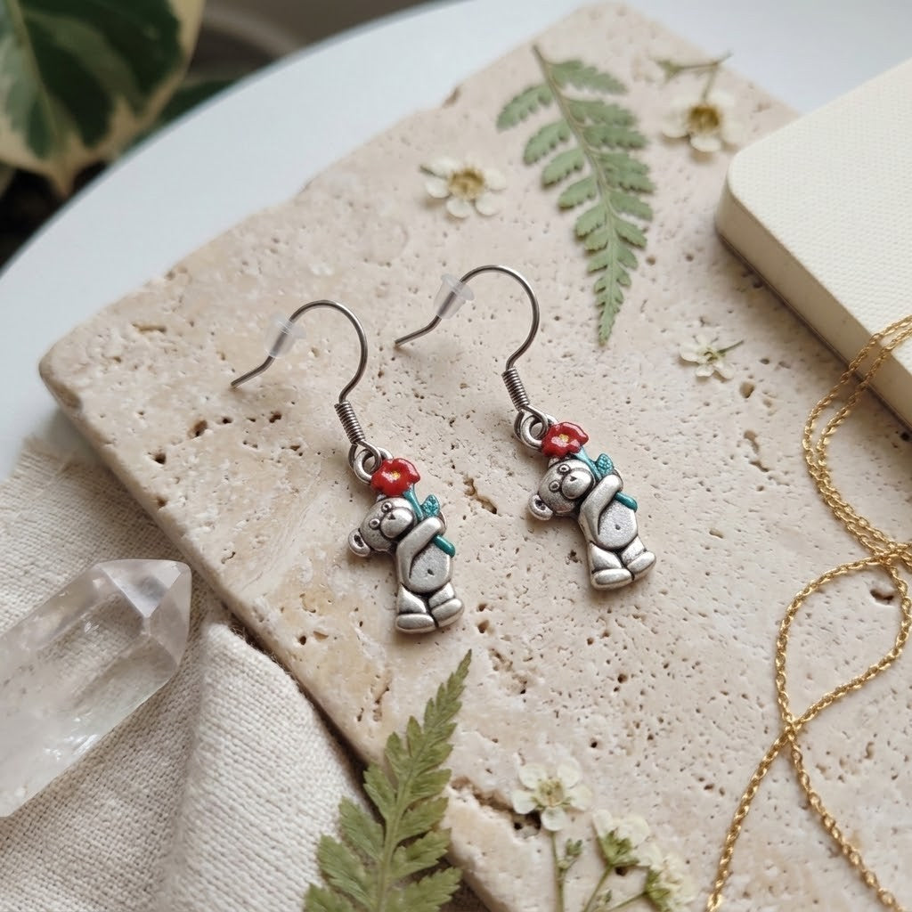 A pair of silver-toned dangle earrings with a small bear charm holding a red heart and a green scarf, displayed on a textured stone slab with botanical and jewelry props.
