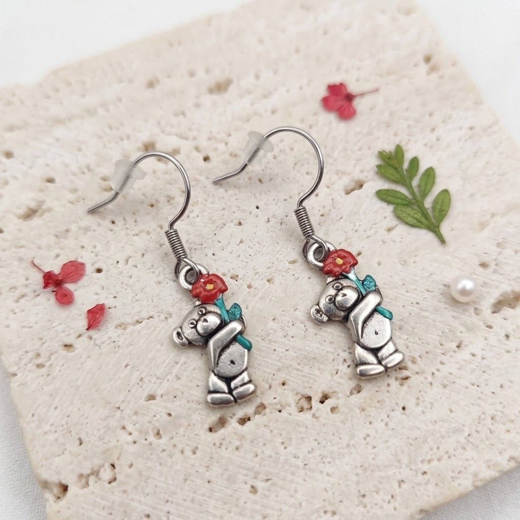 A pair of silver-toned dangle earrings featuring a cute bear character holding a red flower with a teal bow, displayed on a textured beige stone tile with small decorative floral and leaf props.