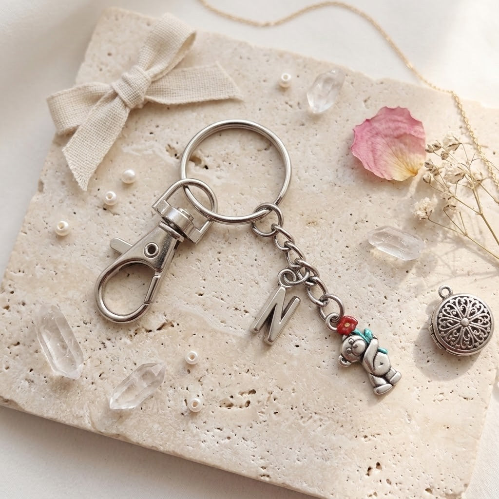 A silver-toned metal keychain with a lobster clasp, a round key ring, a hanging letter charm, and a small bear charm with red and teal accents, styled on a textured beige stone tile with a ribbon, pearls, crystals, and dried flowers.