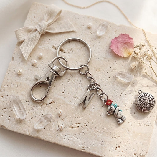 A silver-toned metal keychain with a lobster clasp, a round key ring, a hanging letter charm, and a small bear charm with red and teal accents, styled on a textured beige stone tile with a ribbon, pearls, crystals, and dried flowers.