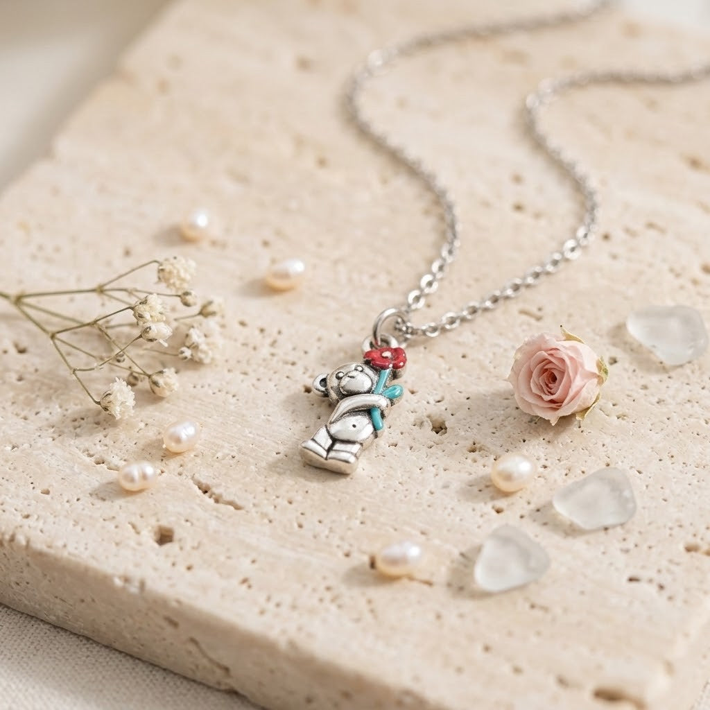 A silver-toned necklace with a teddy bear pendant holding a red and blue flower, styled on a textured beige stone slab with small pearls, dried flowers, and a pink rose.