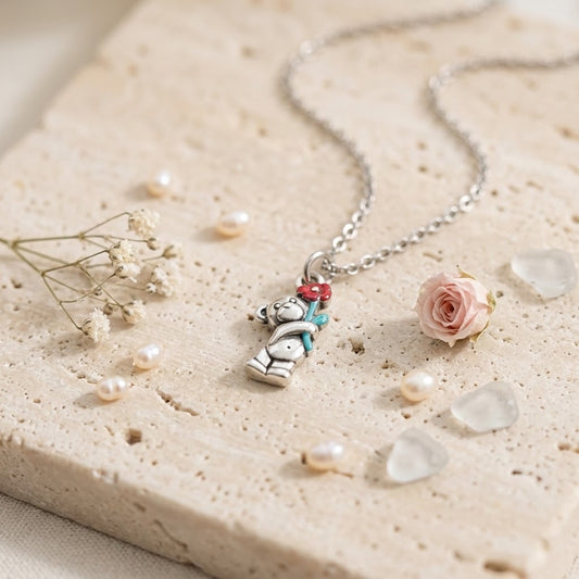 A silver-toned necklace with a teddy bear pendant holding a red and blue flower, styled on a textured beige stone slab with small pearls, dried flowers, and a pink rose.